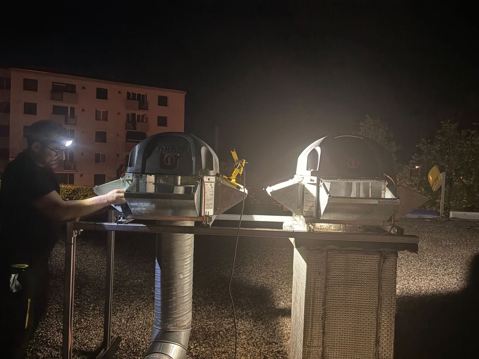 Technicien à la lampe frontale nettoyant deux tourelles d'extraction sur un toit, de nuit